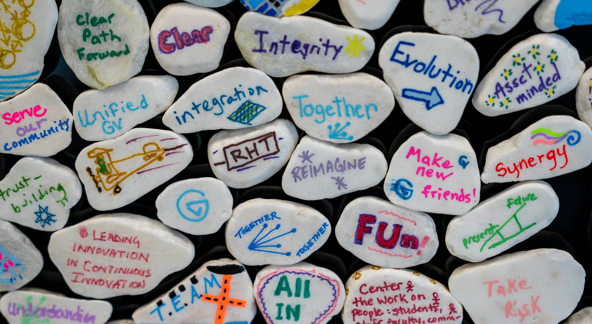 Small white rocks individually decorated with ideas for Reach Higher Together.